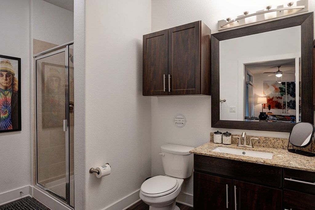 Modern bathroom at The Reserve at Arboretum Apartment Homes with dark wood cabinets, granite countertop, and a large mirror. Glass shower door on the left, vibrant painting on the wall. Cozy ambiance.
