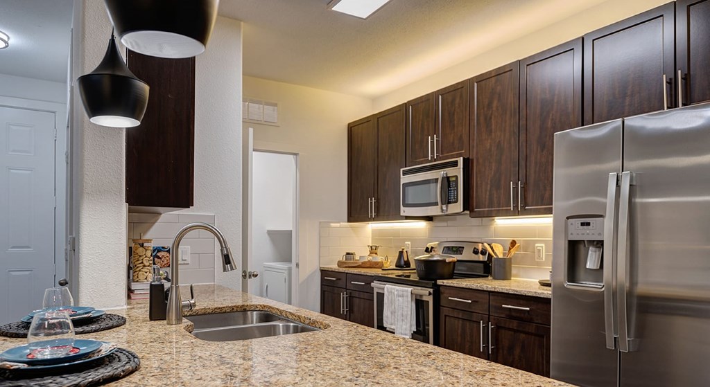 Modern kitchen here at The Reserve at Arboretum Apartment Homes with warm lighting, featuring dark wood cabinets, stainless steel appliances, a granite countertop, and two pendant lights.