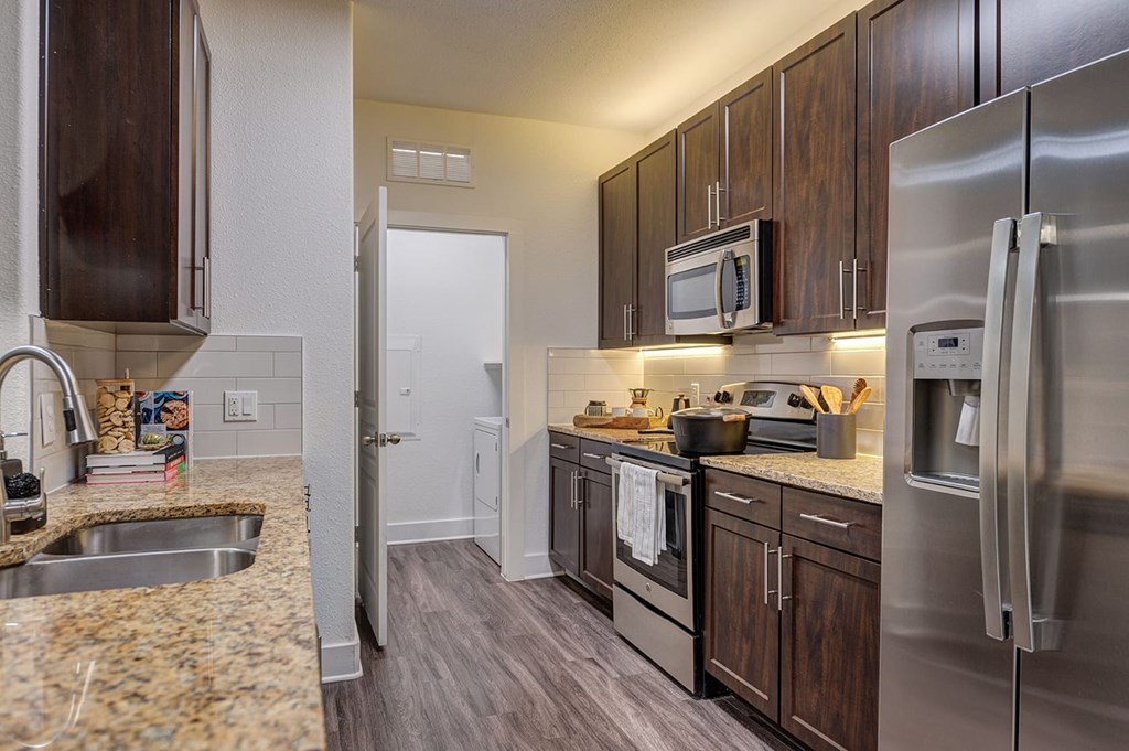 Modern kitchen here at The Reserve at Arboretum Apartment Homes with dark wood cabinets, stainless steel appliances, and granite countertops. A narrow space leads to a laundry area, creating a cozy ambiance.