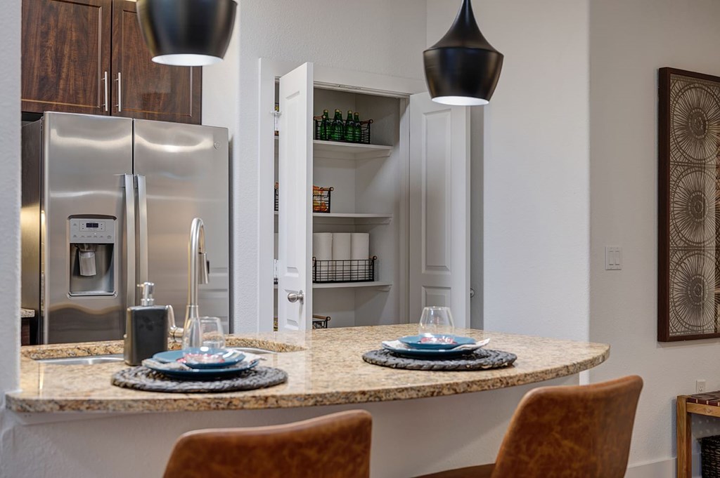 A modern kitchen here at The Reserve at Arboretum Apartment Homes features a granite countertop with two place settings, leather chairs, pendant lights, a stainless steel fridge, and an open pantry.