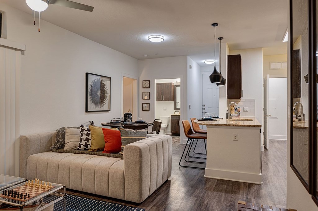 Modern living room here at The Reserve at Arboretum Apartment Homes with a beige sofa adorned with colorful cushions, a chess board, and wall art. Kitchen with pendant lights and bar stools in the background. Warm, inviting atmosphere.