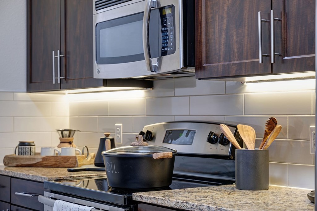 Modern kitchen here at The Reserve at Arboretum Apartment Homes with dark wood cabinets, stainless steel microwave, black stove with a pot, wooden utensils in a holder, and soft under-cabinet lighting.