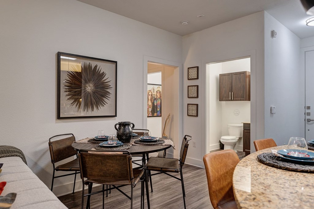 Dining area in modern apartment here at The Reserve at Arboretum Apartment Homes with a round table set for four, dark chairs, and a decorative jug. Art hangs on white walls, creating a cozy atmosphere.