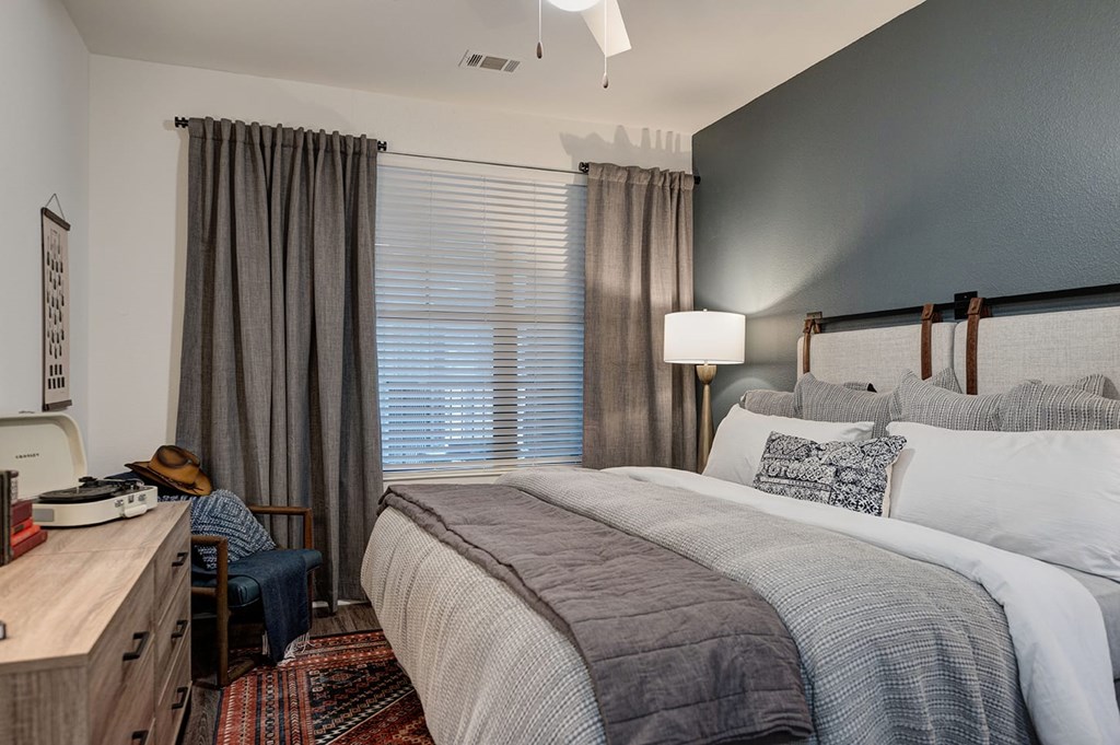 A cozy bedroom at The Reserve at Arboretum Apartment Homes with a neatly made bed featuring gray and white bedding. A lamp illuminates a dark accent wall, while curtains frame a large window.