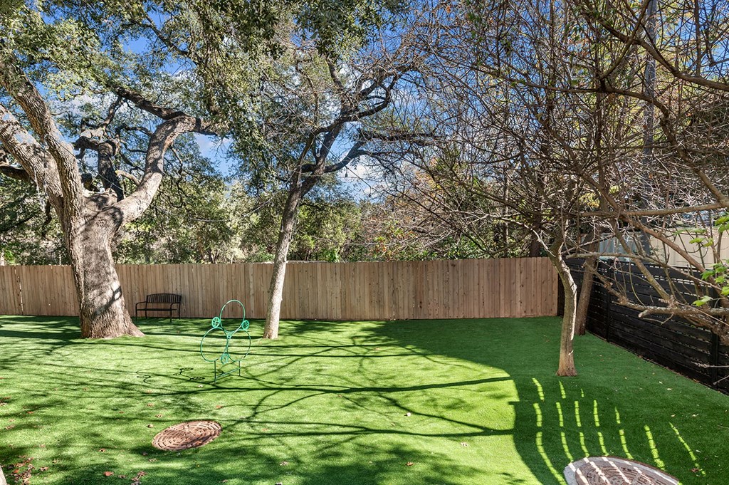 A sunlit backyard here at The Reserve at Arboretum Apartment Homes with artificial grass, large twisting trees, and a small bench near a wooden fence. A green garden decoration is in the center.