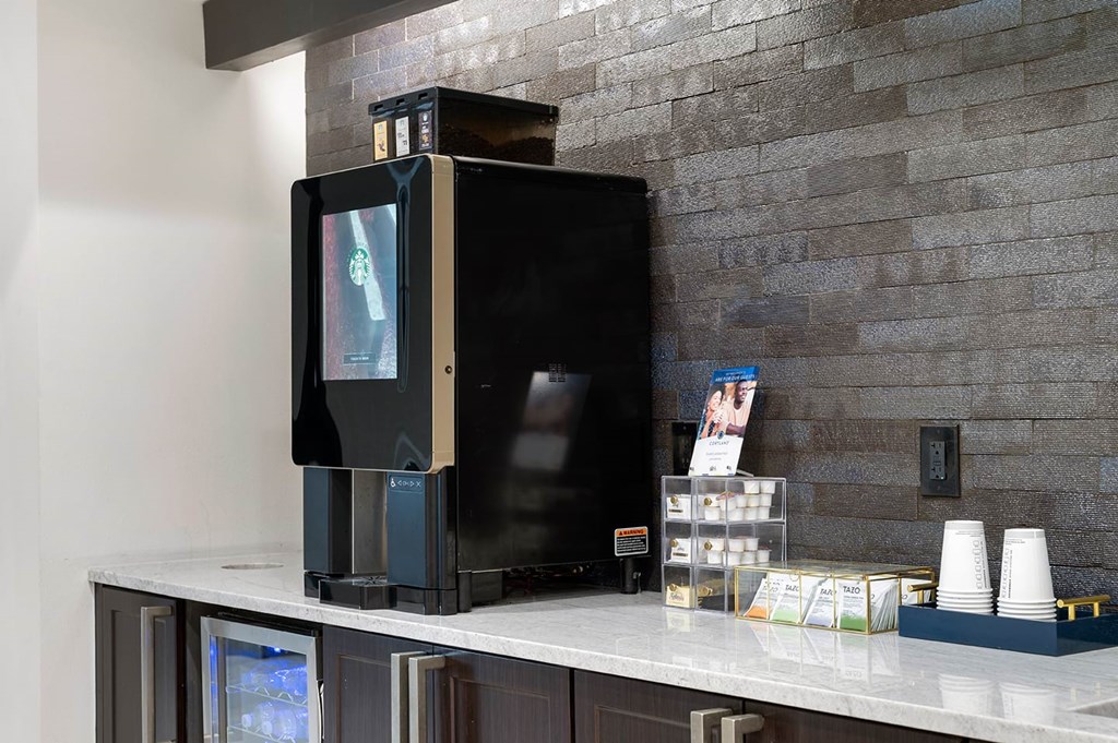 A sleek, black coffee machine sits on a marble counter against a textured gray wall here at The Reserve at Arboretum Apartment Homes. Nearby are stacked paper cups and a small display of creamers, creating a modern, inviting coffee station.