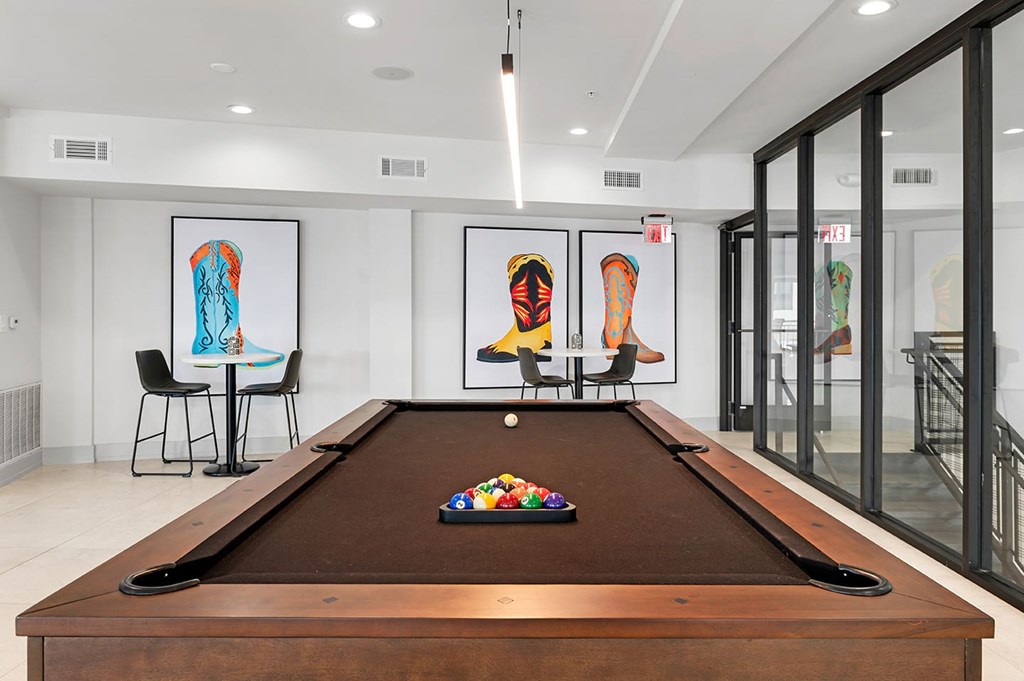 Modern game room here at The Reserve at Arboretum Apartment Homes with a brown pool table centered, surrounded by vibrant cowboy boot artwork on white walls. Black chairs and a table are nearby.