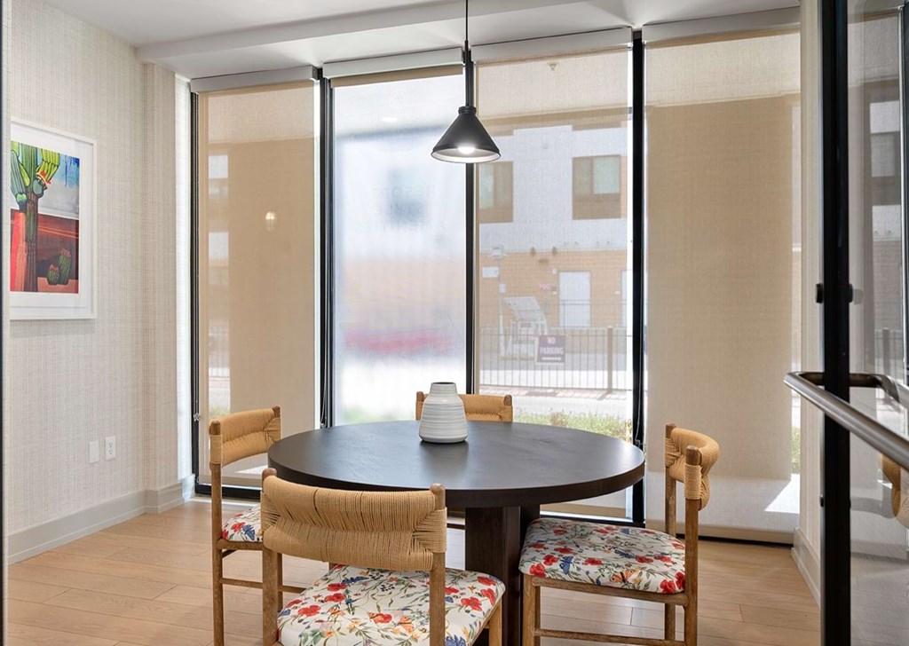 A modern dining area here at The Reserve at Arboretum Apartment Homes with a round black table, a white vase centerpiece, and four chairs with floral cushions. Large windows diffuse natural light.
