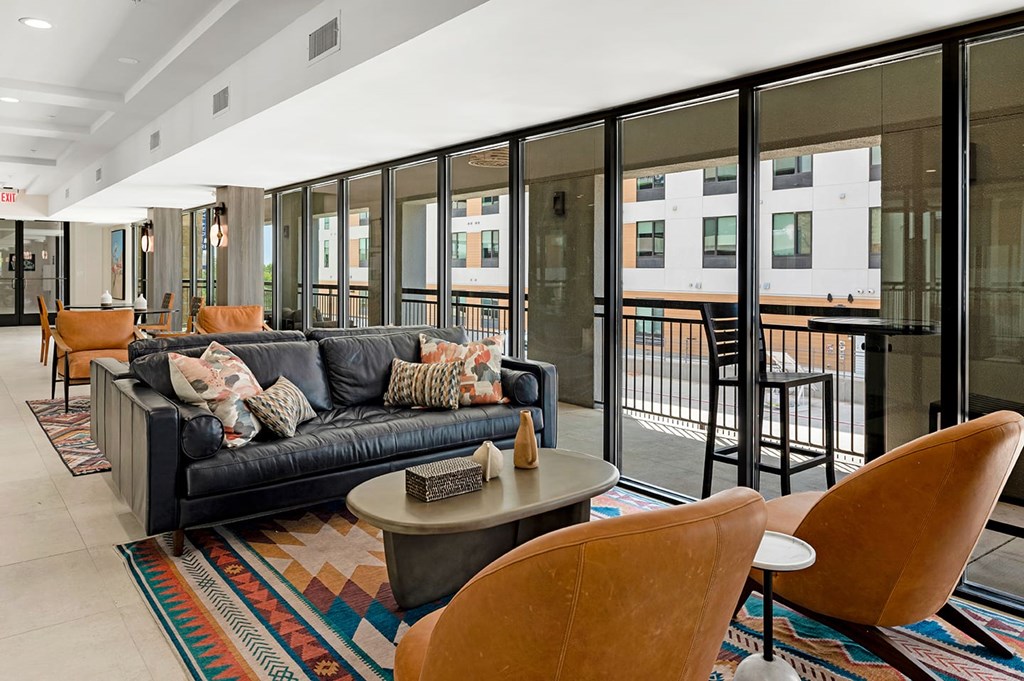 Modern lounge here at The Reserve at Arboretum Apartment Homes with a black leather sofa adorned with patterned pillows, flanked by brown armchairs. Large windows offer urban views, creating a cozy yet sleek ambiance.