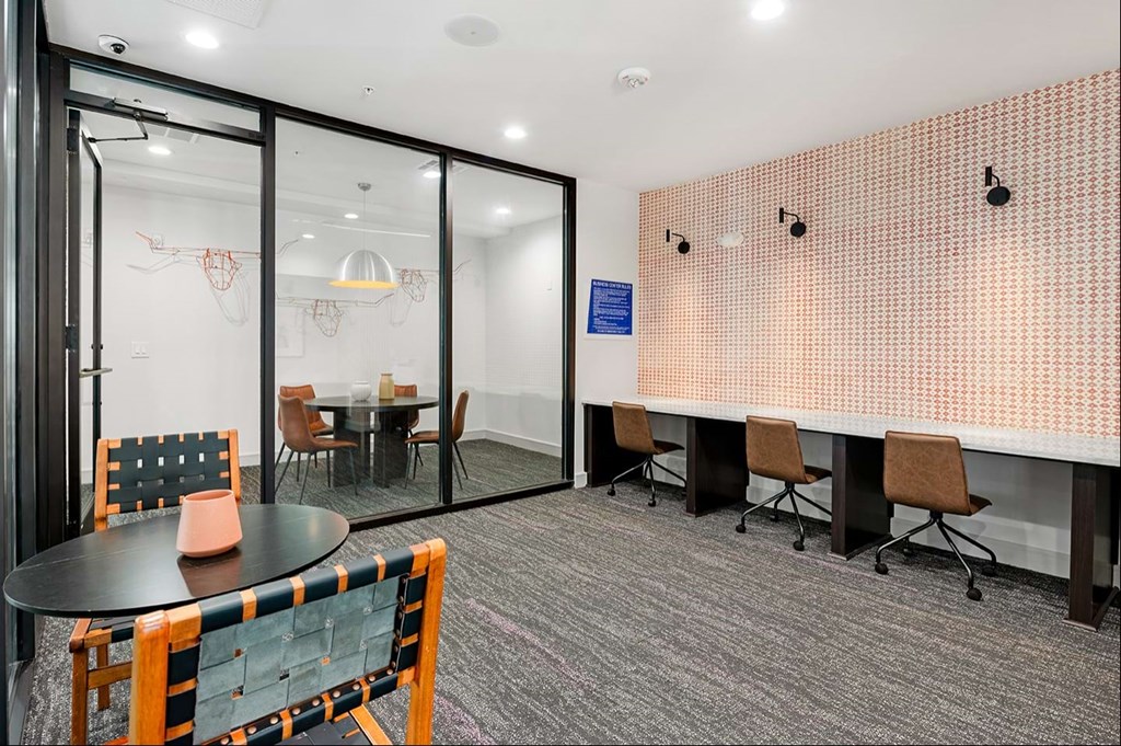 Modern office with neutral tones here at The Reserve at Arboretum Apartment Homes. Features a small meeting room with a glass wall, a sitting area with round table, and three workstations.