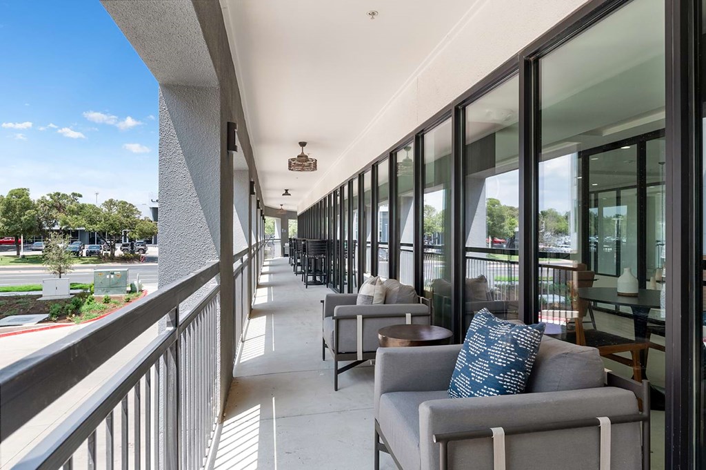 Spacious modern balcony here at The Reserve at Arboretum Apartment Homes with glass doors, gray armchairs, and blue-patterned pillows. Overlooks a sunny street and greenery, conveying relaxation.
