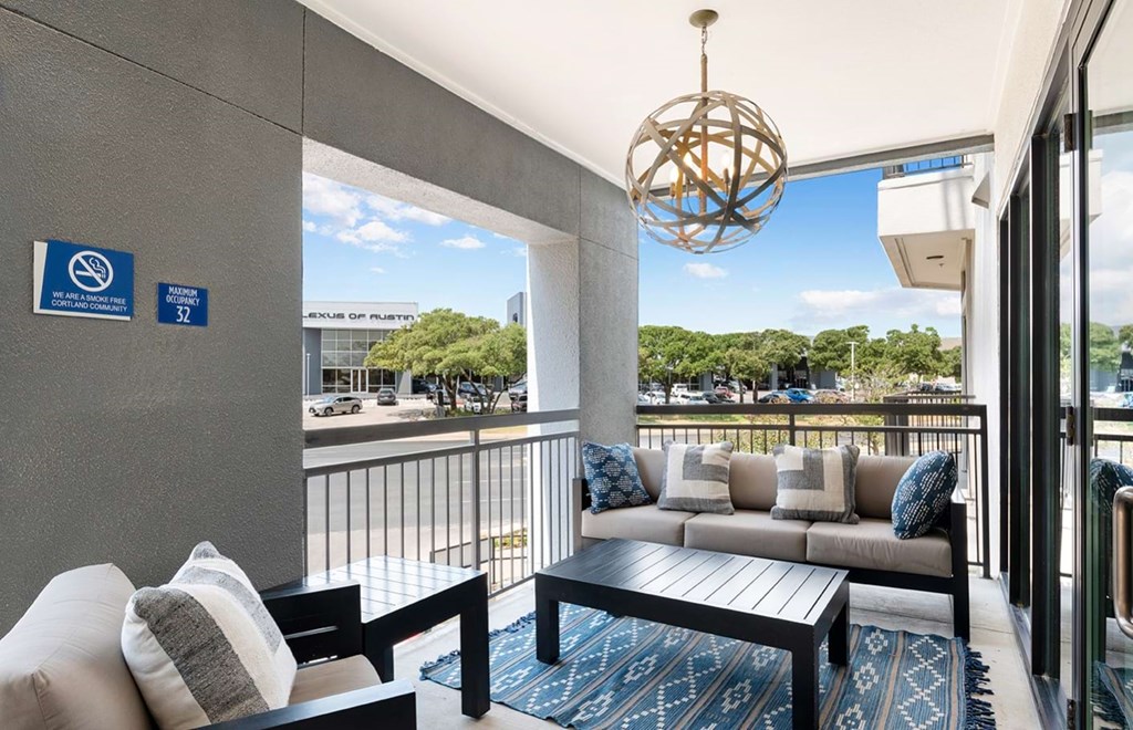 Outdoor patio here at The Reserve at Arboretum Apartment Homes with stylish seating, blue and white cushions, and geometric hanging light. Calm, modern vibe overlooking trees and parking area.
