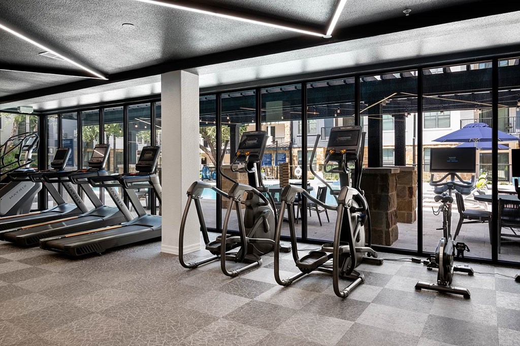 Modern gym interior here at The Reserve at Arboretum Apartment Homes with treadmills and exercise bikes facing large windows. The view outside shows a pool area with umbrellas, creating an inviting atmosphere.