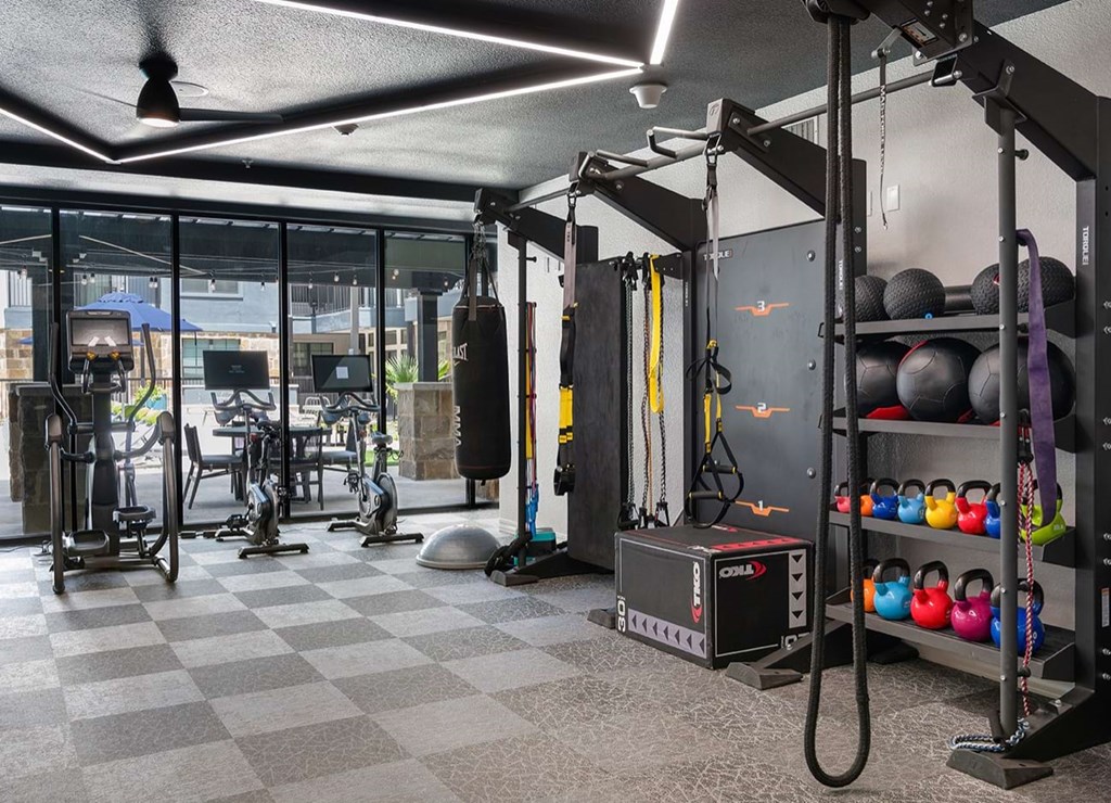 Modern gym here at The Reserve at Arboretum Apartment Homes with exercise equipment, including bikes, a punching bag, kettlebells, and fitness bands. Bright lighting and large windows create an energetic atmosphere.