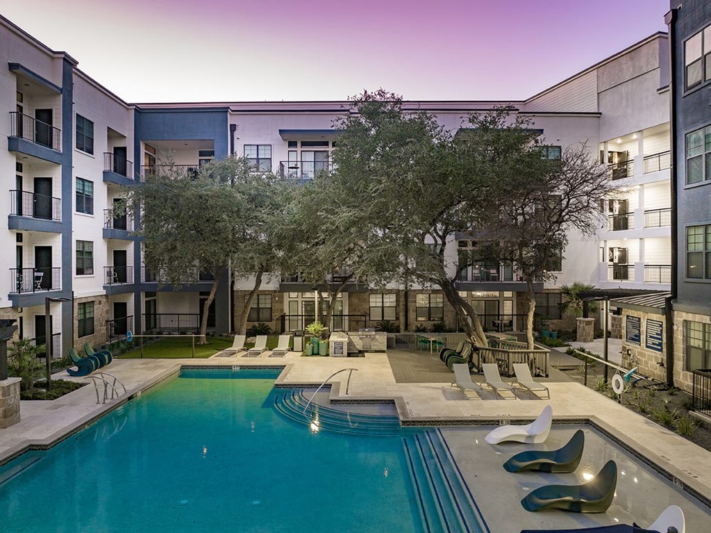 Modern courtyard here at The Reserve at Arboretum Apartment Homes at dusk with a lit swimming pool, surrounded by deck chairs and trees. The sky is a soft gradient of purple and pink.