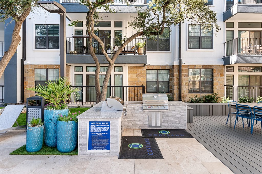 Outdoor patio with a gas grill station here at The Reserve at Arboretum Apartment Homes, surrounded by blue planters and trees. Modern apartments in the background. Clean, inviting atmosphere.