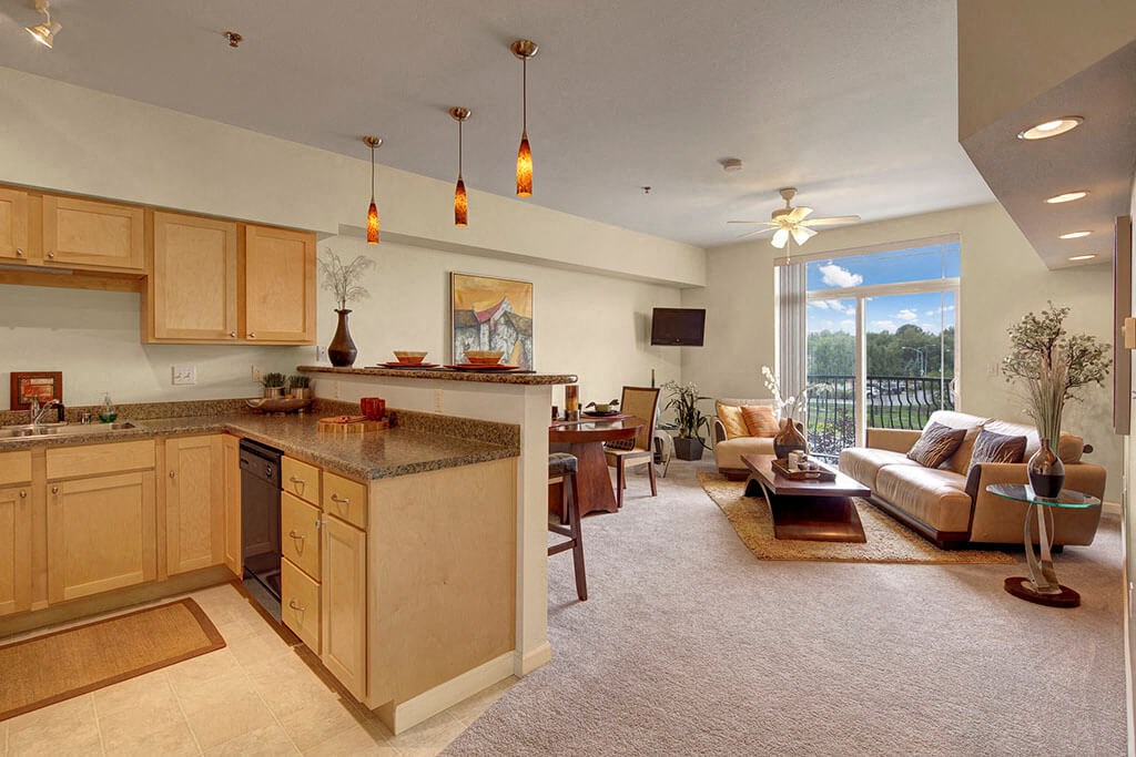 Spacious open-plan living room and kitchen here at Park Plaza 2 Apartments. Light wood cabinets, pendant lights, cozy seating, large window with greenery view, warm, inviting atmosphere.