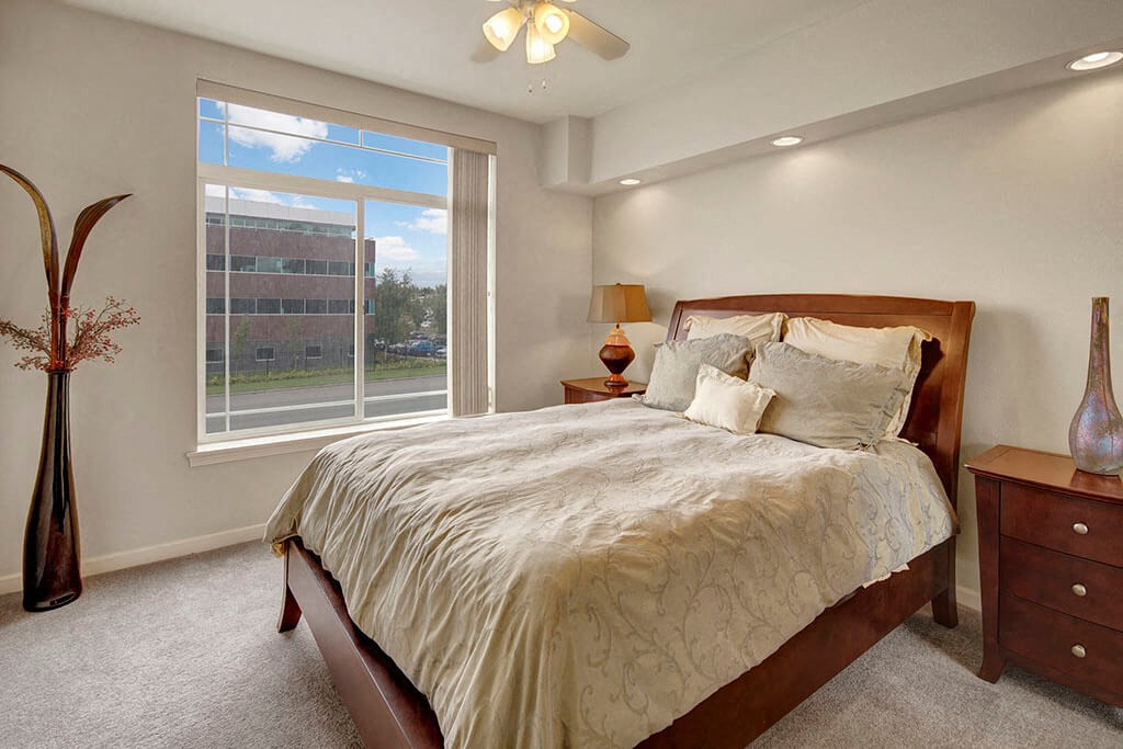 Park Plaza II Apartments - Bedroom Modern bedroom here at Park Plaza 2 Apartments with a large window view, featuring a wooden bed with beige and gray bedding, side tables, a tall vase, and a warm, inviting ambiance.