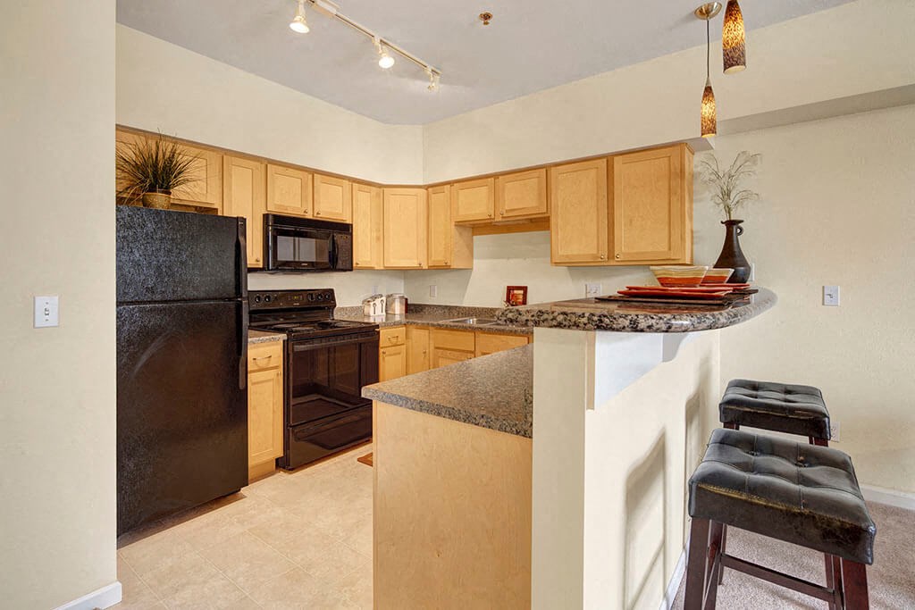 Park Plaza II Apartments - Kitchen Modern kitchen here at Park Plaza 2 Apartments with light wood cabinets and black appliances, including a fridge and stove. Granite countertops, pendant lighting, and bar stools add elegance.