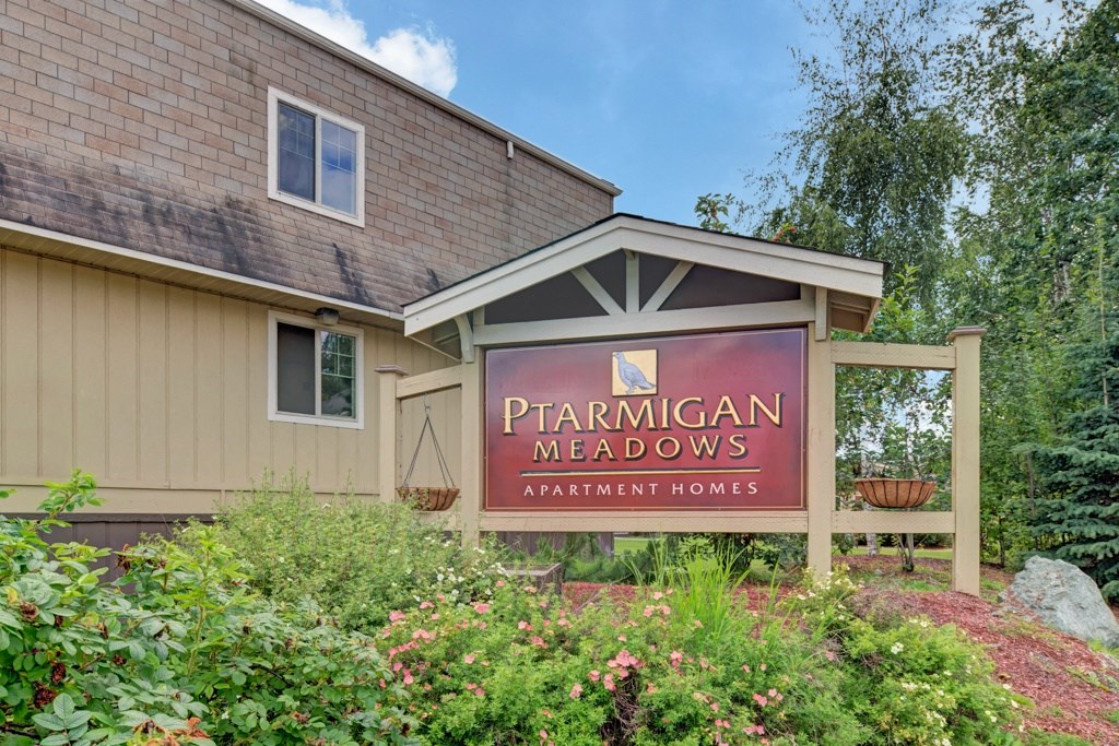 Sign reading "Ptarmigan Meadows Apartment Homes," surrounded by lush greenery and flowers, sits in front of a beige building under a partly cloudy sky.