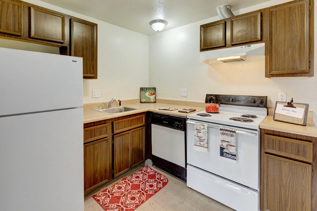 Compact kitchen here at Ptarmigan Meadows with wood cabinets, white appliances, and a red kettle on the stove. A red patterned rug adds warmth. Decor includes coffee-themed towels.