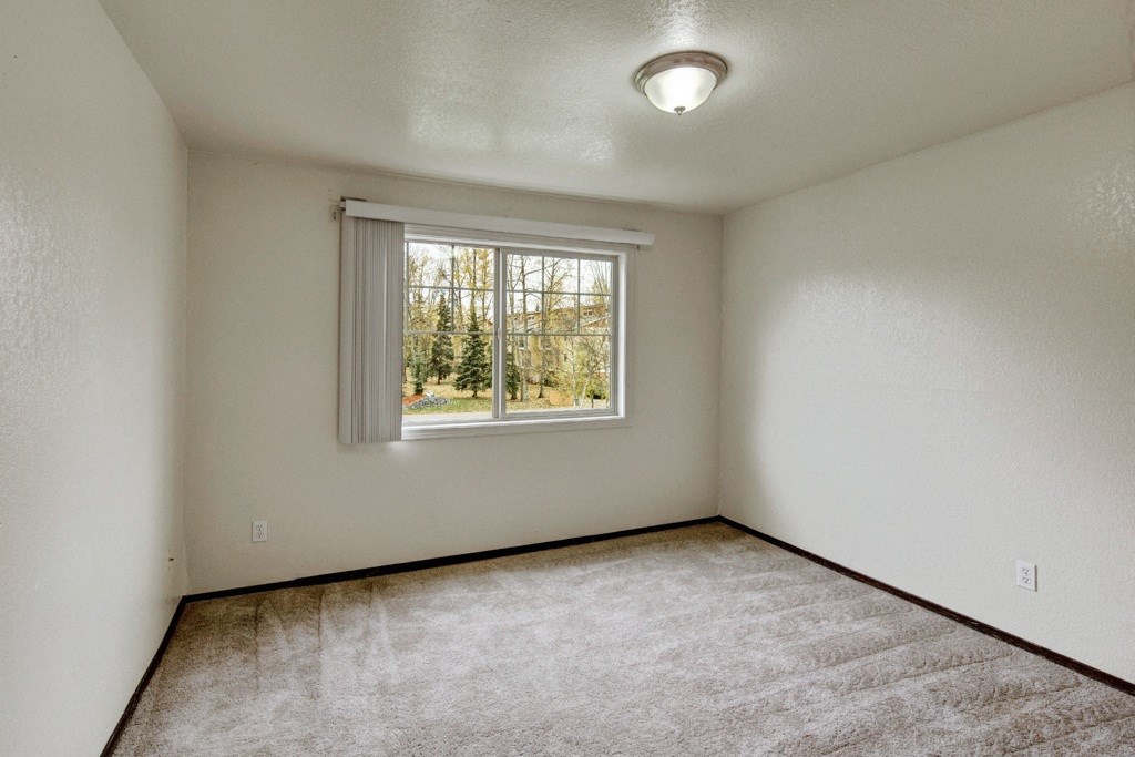 Small, empty bedroom here at Ptarmigan Meadows with beige walls and carpet. A single window with vertical blinds shows a view of trees outside. Ceiling light fixture adds a calm tone.