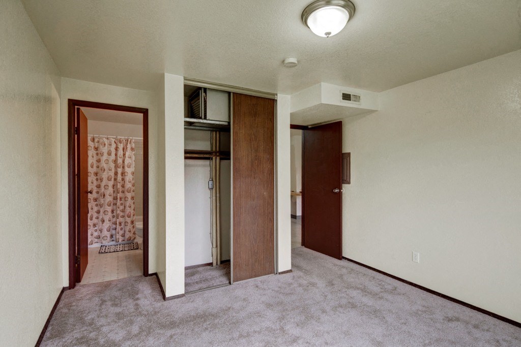 Empty bedroom here at Ptarmigan Meadows with beige carpet and white walls. An open closet with wooden sliding doors is on the right, and a bathroom visible through a door on the left.