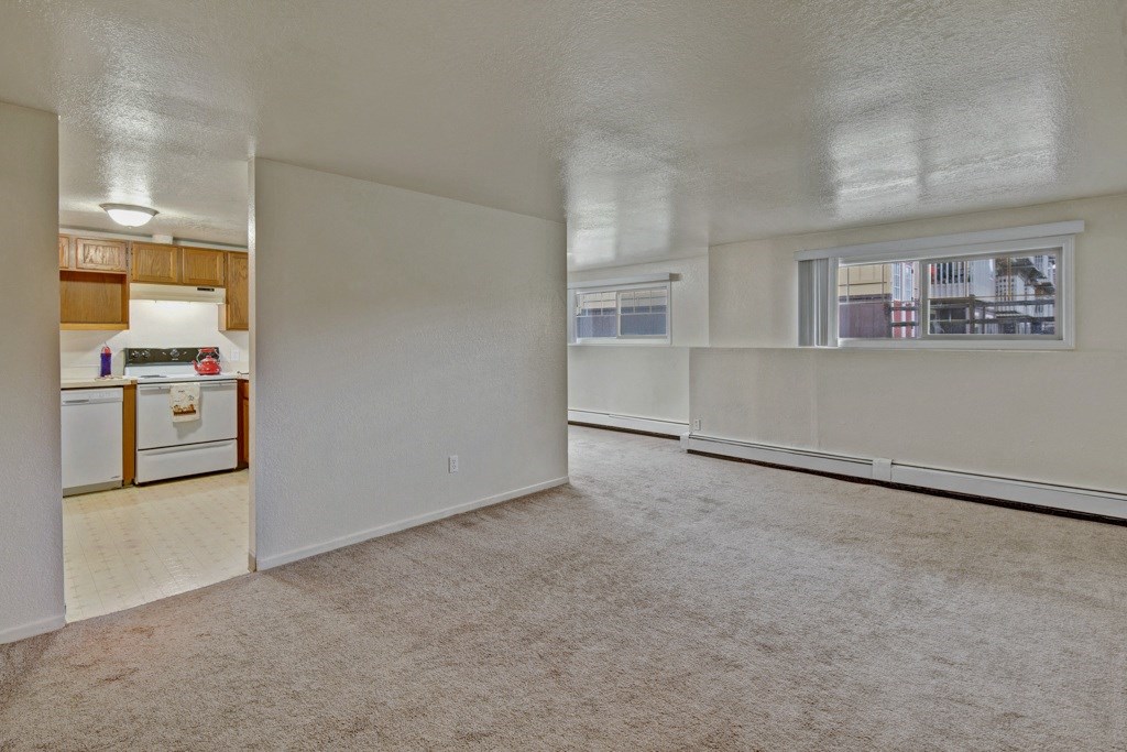 Spacious unfurnished room here at Ptarmigan Meadows with beige carpet and white walls. Adjacent kitchen has wooden cabinets, stove, and dishwasher. Large window offers natural light.