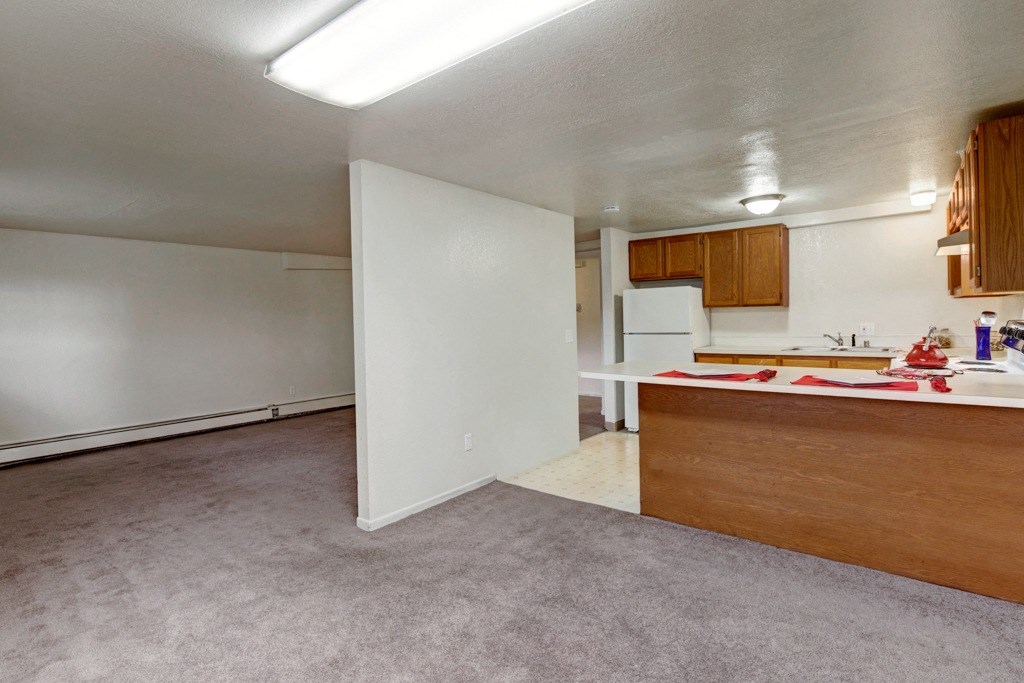 Open-plan room here at Ptarmigan Meadows with beige walls, carpeted living area, and a kitchen featuring wooden cabinets and a white fridge. A cozy, neutral space.