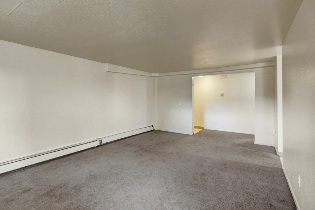 Empty living room here at Ptarmigan Meadows with beige carpet and off-white walls. Open doorway leads to a small, lighted area. Sparse, calm atmosphere with a minimalist feel.