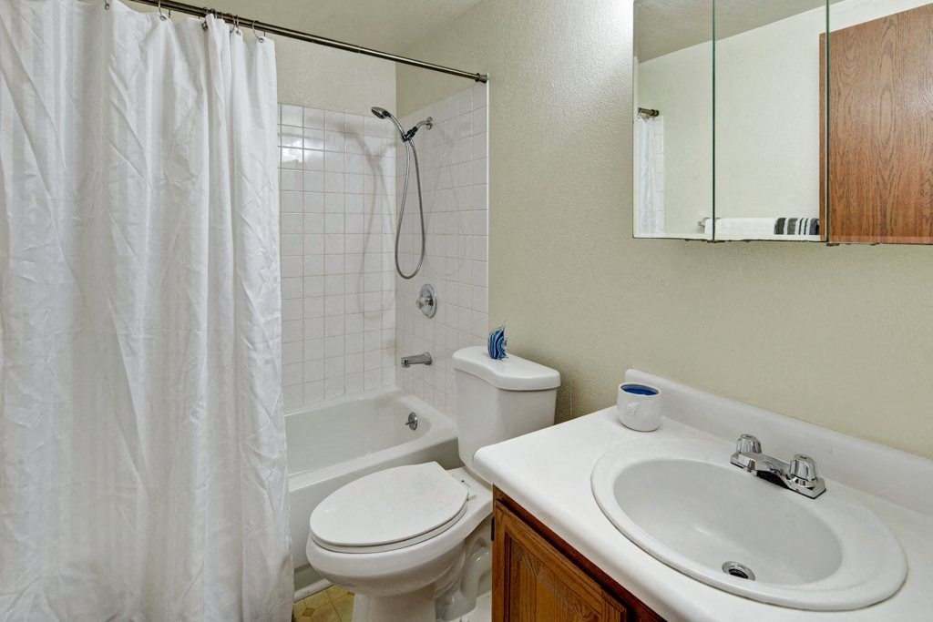 Small bathroom here at Ptarmigan Meadows with white tiled bathtub and shower, white curtain, toilet, and sink with wooden cabinet. Mirror cabinet and simple, clean decor.