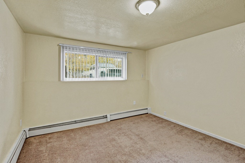 Empty beige bedroom here at Ptarmigan Meadows with brown carpet, a ceiling light, and a window with blinds overlooking a parked car. The space feels calm and neutral.