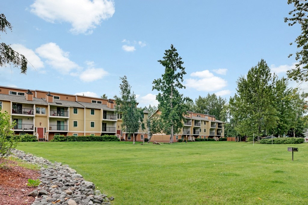 Ptarmigan Meadows Apartment complex with yellow-brown buildings, surrounded by trees and an expansive grassy area. Bright, sunny day with a few clouds in the sky.