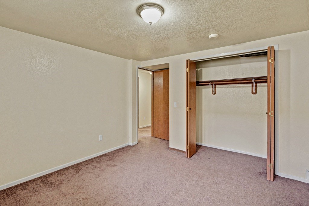 An empty bedroom here at Ptarmigan Meadows with beige walls and a brown carpet. Open closet with wooden doors and a hanging rod. Ceiling light illuminates the space, creating a simple, neutral tone.