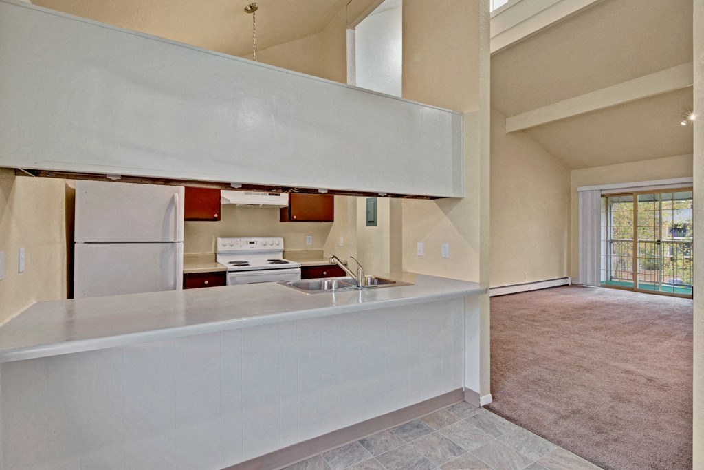 Open-plan kitchen and living room here at Ptarmigan Meadows with neutral tones. Kitchen features white appliances and brown cabinets. Living area has large windows and balcony access.