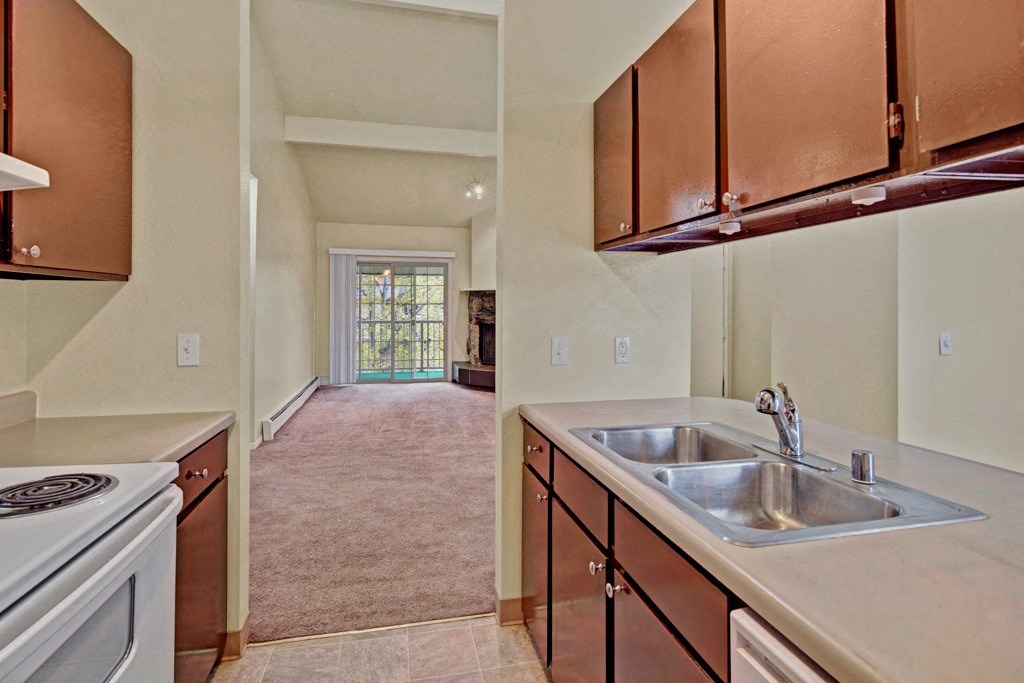Narrow kitchen here at Ptarmigan Meadows with dark wood cabinets, double stainless steel sink, and white stove. Leads to a carpeted living room with large glass doors.
