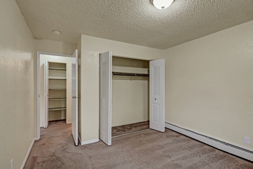 Small empty bedroom here at Ptarmigan Meadows with beige walls and carpet, featuring an open closet with louvered doors and a doorway leading to a shelf-lined closet, lit by a ceiling light.