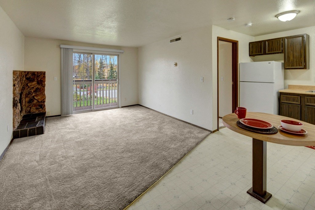 Cozy apartment interior here at Ptarmigan Meadows with beige carpet, sliding glass doors, small brick fireplace, and compact kitchen. A round table holds red dinnerware, creating a warm, inviting atmosphere.