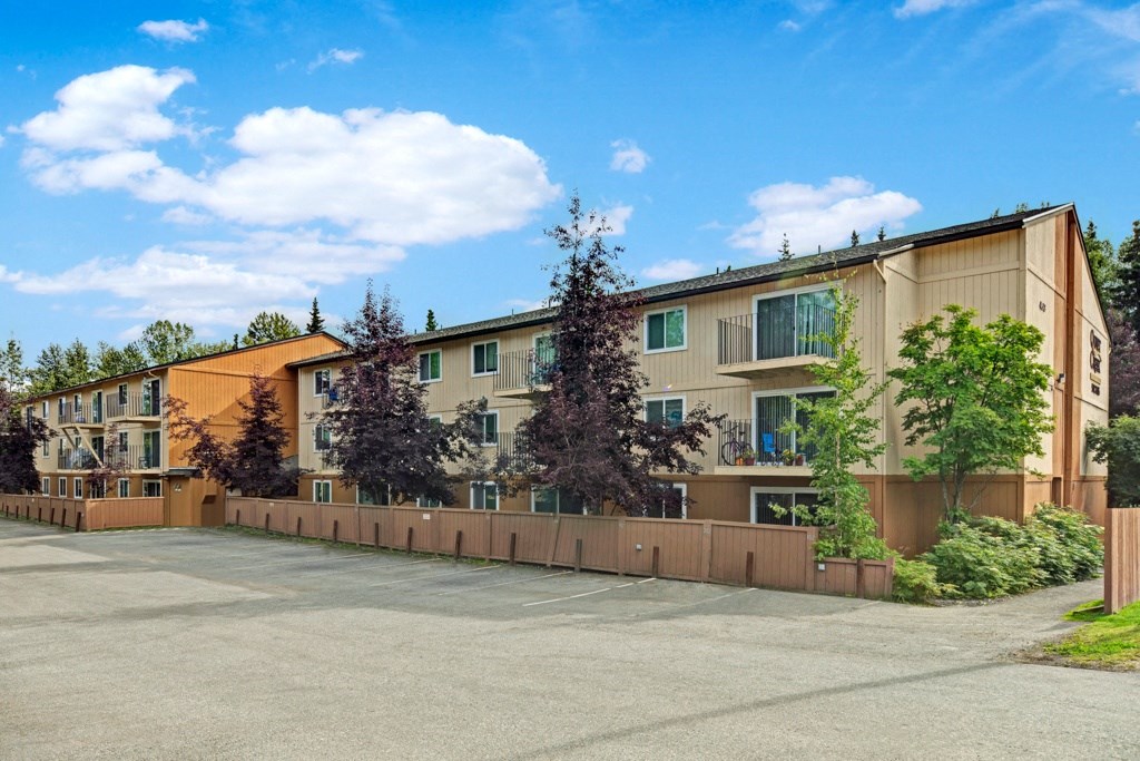 Three-story beige Quiet Creek Apartment building with brown trim, surrounded by trees and enclosed by a wooden fence, under a bright blue sky with scattered clouds.