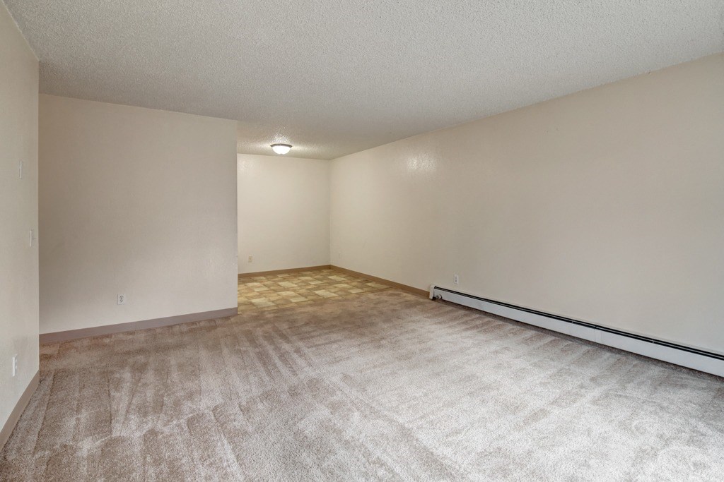 Empty living room here at Quiet Creek Apartments with beige carpet flooring, light cream walls, and a baseboard heater on the right. A lit ceiling light is visible in the back corner. Minimal and clean atmosphere.