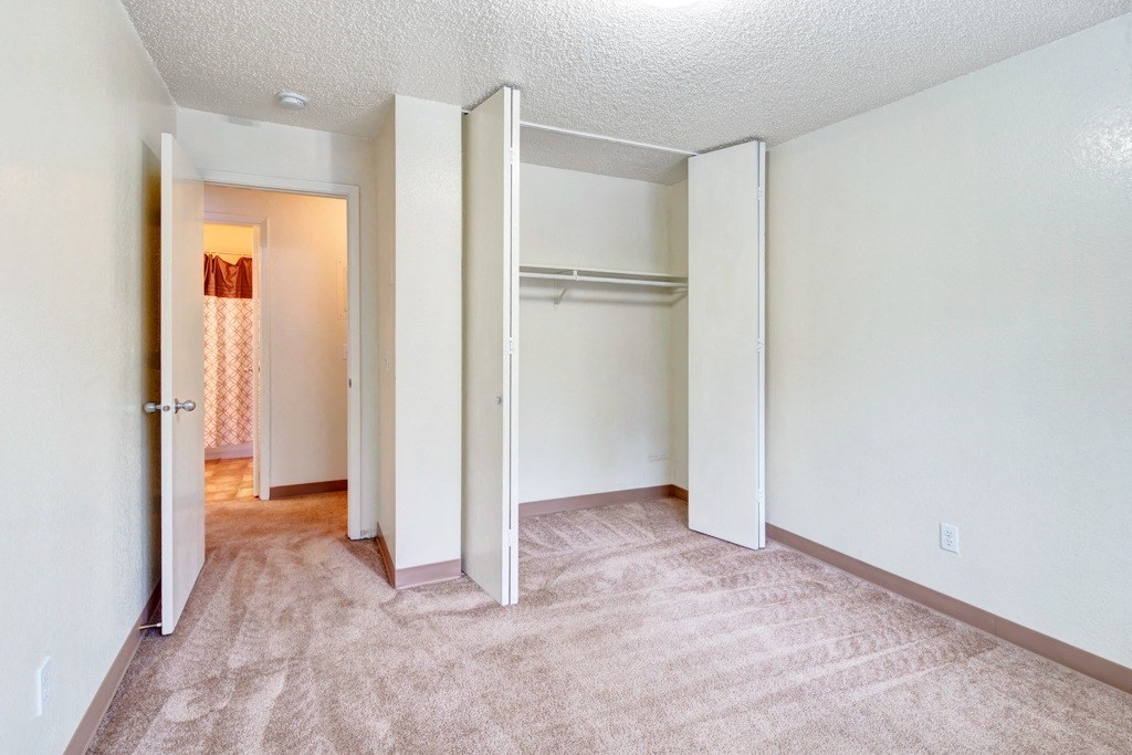 Empty bedroom here at Quiet Creek Apartments with beige carpet and white walls, featuring an open closet with shelves. A door leads to a lit hallway with a patterned shower curtain.