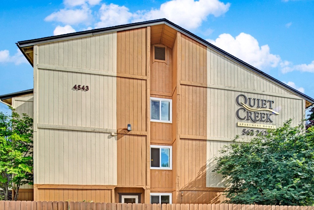 Apartment building with wood paneling in beige and tan. "Quiet Creek Apartment Homes" sign on the right. Trees and a wooden fence in the foreground. Blue sky above.