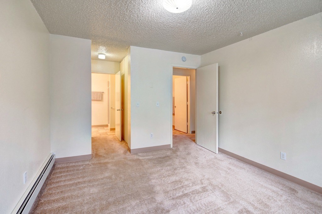 Empty bedroom here at Quiet Creek Apartments with beige carpet and off-white walls. An open door reveals a lit hallway. The room is minimalistic and calm, evoking a neutral tone.