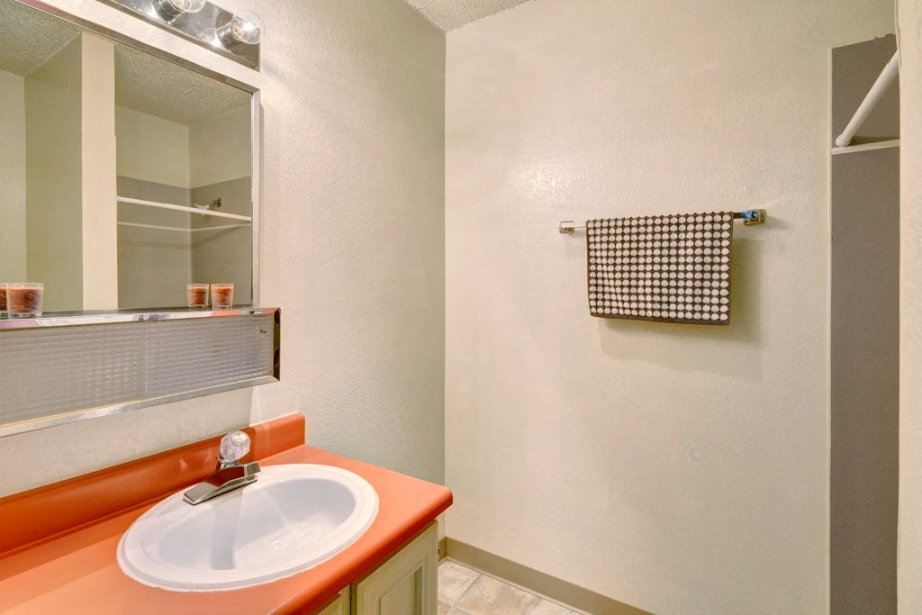 A small bathroom here at Quiet Creek Apartments with a coral countertop sink, a lit mirror above, and a patterned towel on a rack. Neutral tones create a clean, simple feel.