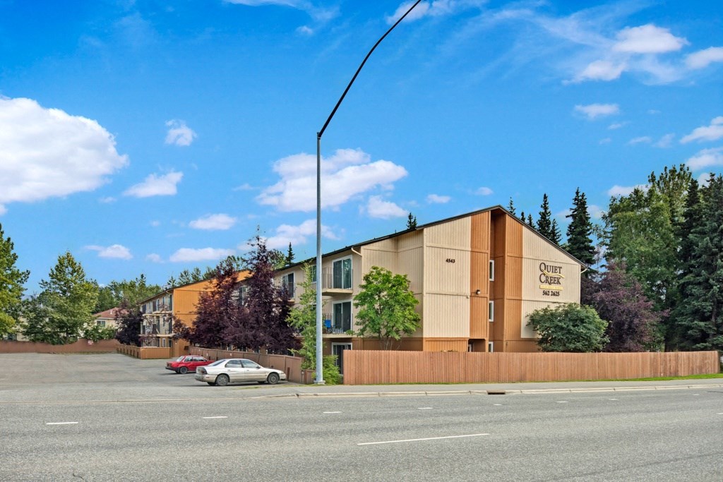 Three-story apartment building with "Quiet Creek" sign, surrounded by trees, under a blue sky with clouds. Two cars parked in front on a paved lot.