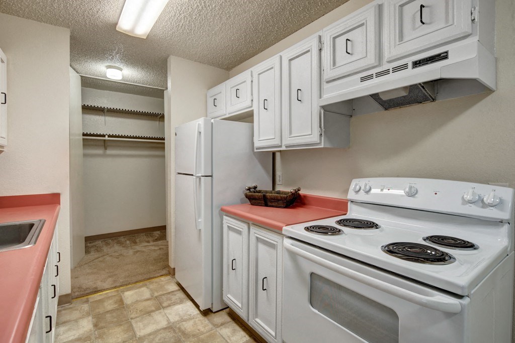 Narrow kitchen here at Quiet Creek Apartments with white cabinets, red countertops, and a white stove. A white refrigerator stands beside an open pantry with empty shelves. Bright lighting.