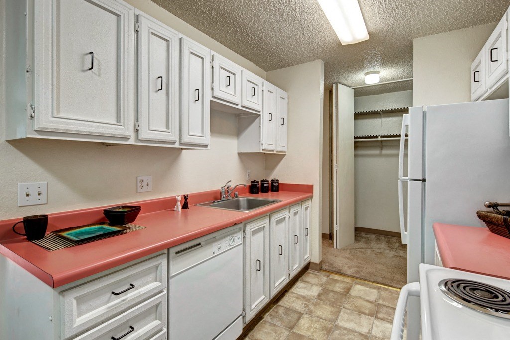 Narrow kitchen here at Quiet Creek Apartments with coral countertops, white cabinets, stove, and fridge. Bright lighting, clean, organized, with a modern yet cozy feel.