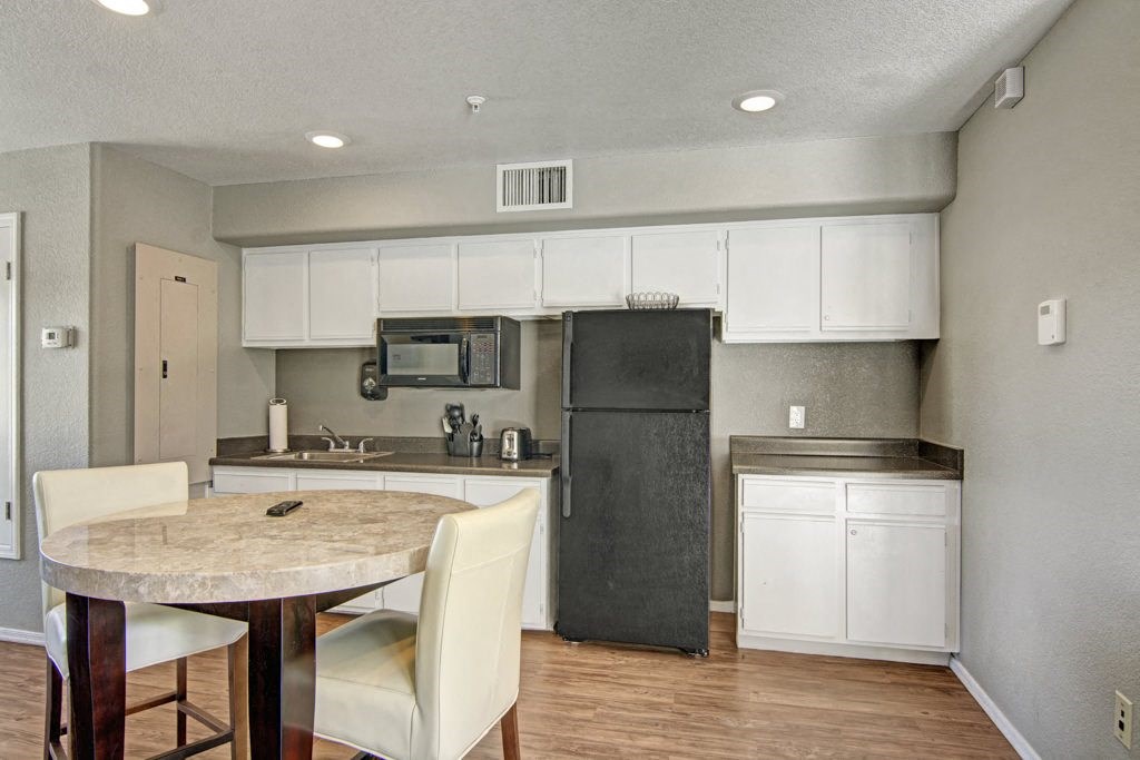Small kitchen here at Reserve at Arrowhead Apartments with white cabinets, black fridge, and appliances on the counter. A round table with beige chairs sits on wood flooring, creating a neutral, cozy atmosphere.