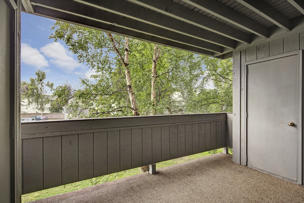Covered balcony here at Russian Jack Apartments with wooden railing, beige carpet, and closed gray door, overlooking lush green trees. Bright sky and distant buildings create a peaceful setting.