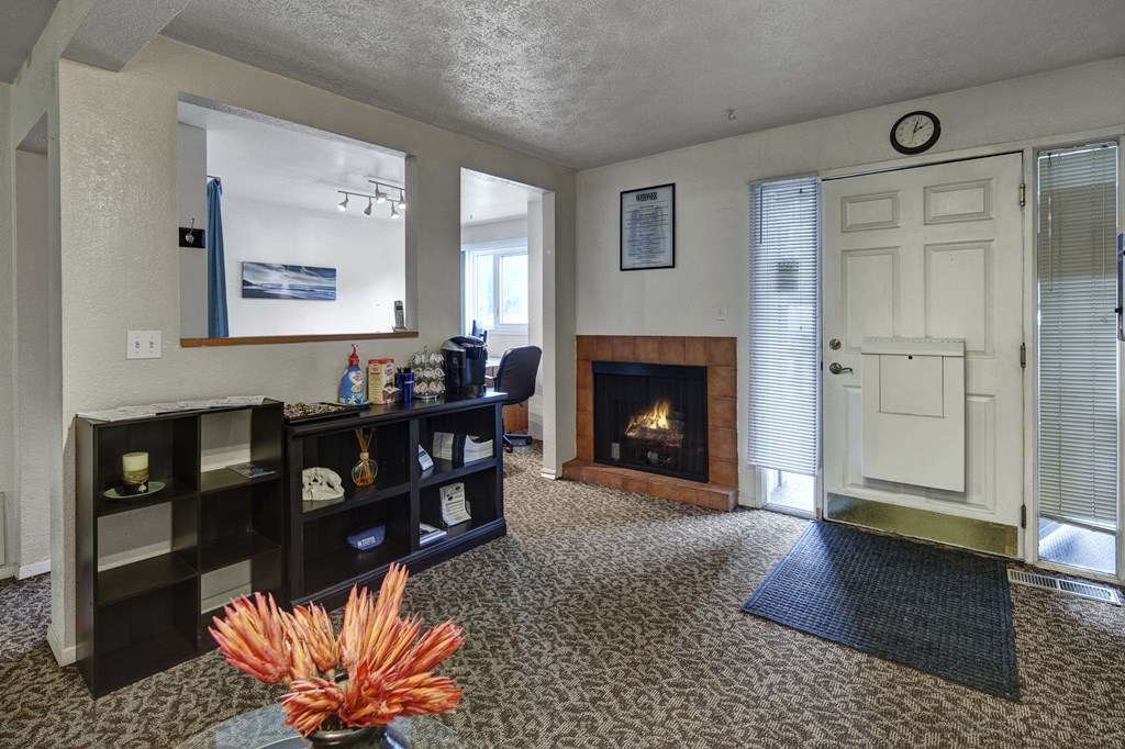 Cozy office entrance here at Russian Jack Apartments with a lit fireplace, shelf with snacks and supplies, and a large clock above a white door flanked by blinds. Warm ambiance.